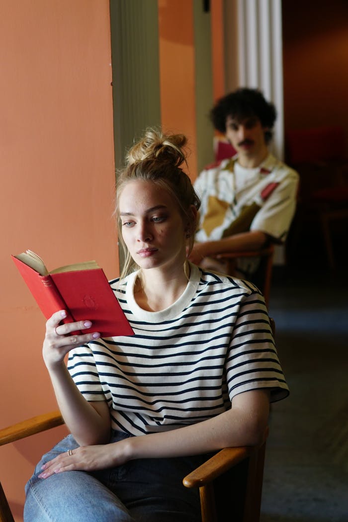 Woman in striped shirt reading a red book, seated indoors with background figure out of focus.