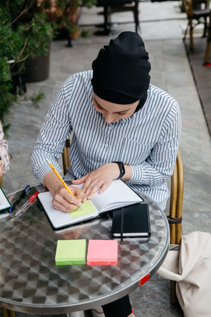 Muslim woman in hijab writing in notebook at outdoor café table. Productive work environment.
